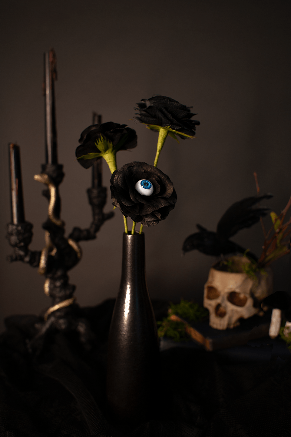 Black paper roses, one with a single realistic blue eye in the center, placed in a black bottle vase, styled on a dark tablecloth with a black candelabra and a skull decor in the background.