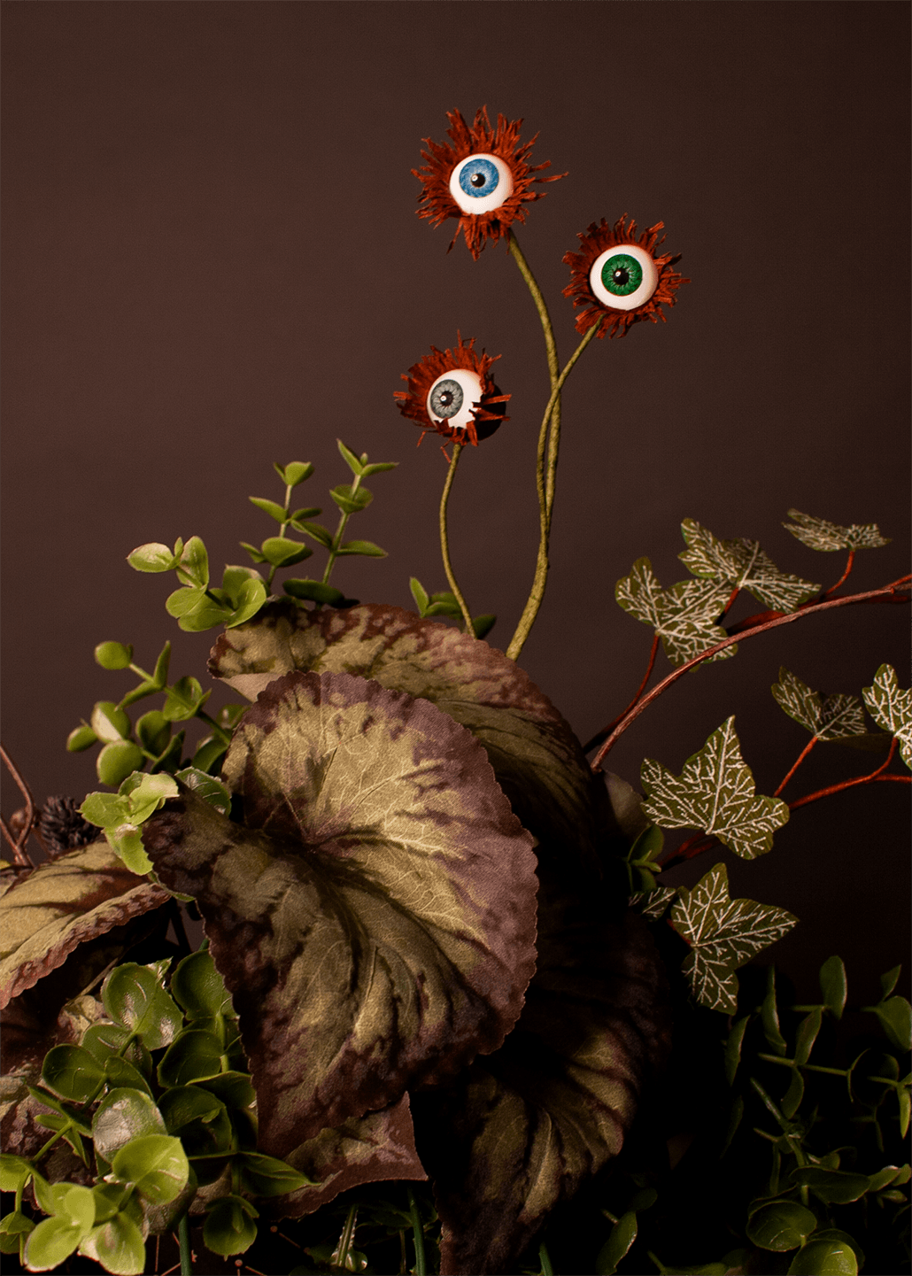 A decorative floral arrangement featuring three artificial eyeball flowers with red spiky centers, green and silver foliage, and large mottled leaves against a dark grey background.
