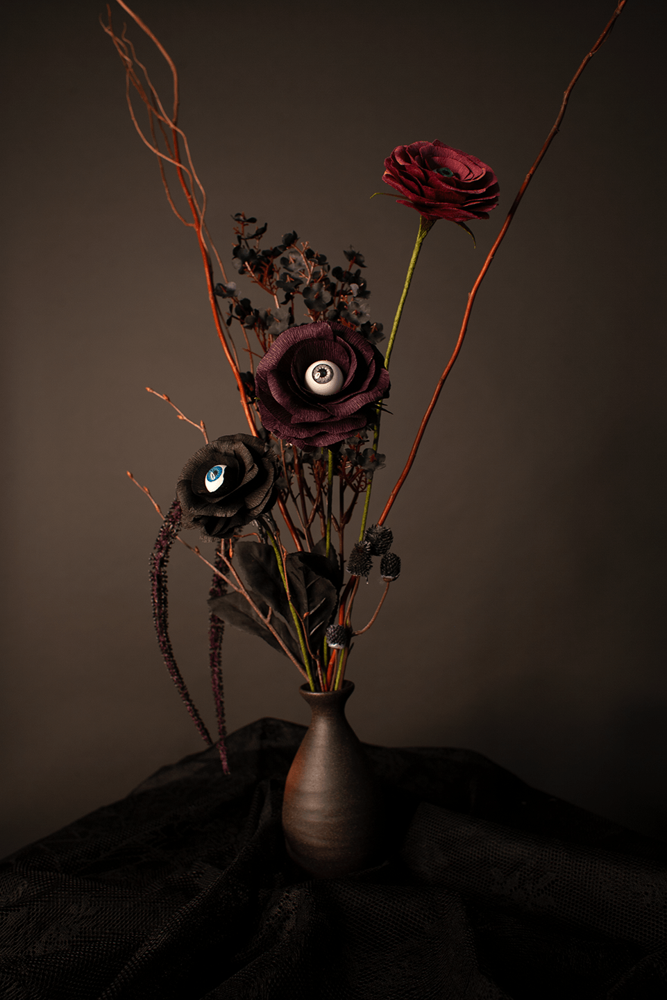 Gothic paper flower arrangement featuring dark red and black roses with realistic eyes, set in a dark ceramic vase on a draped black cloth against a dark brown background.