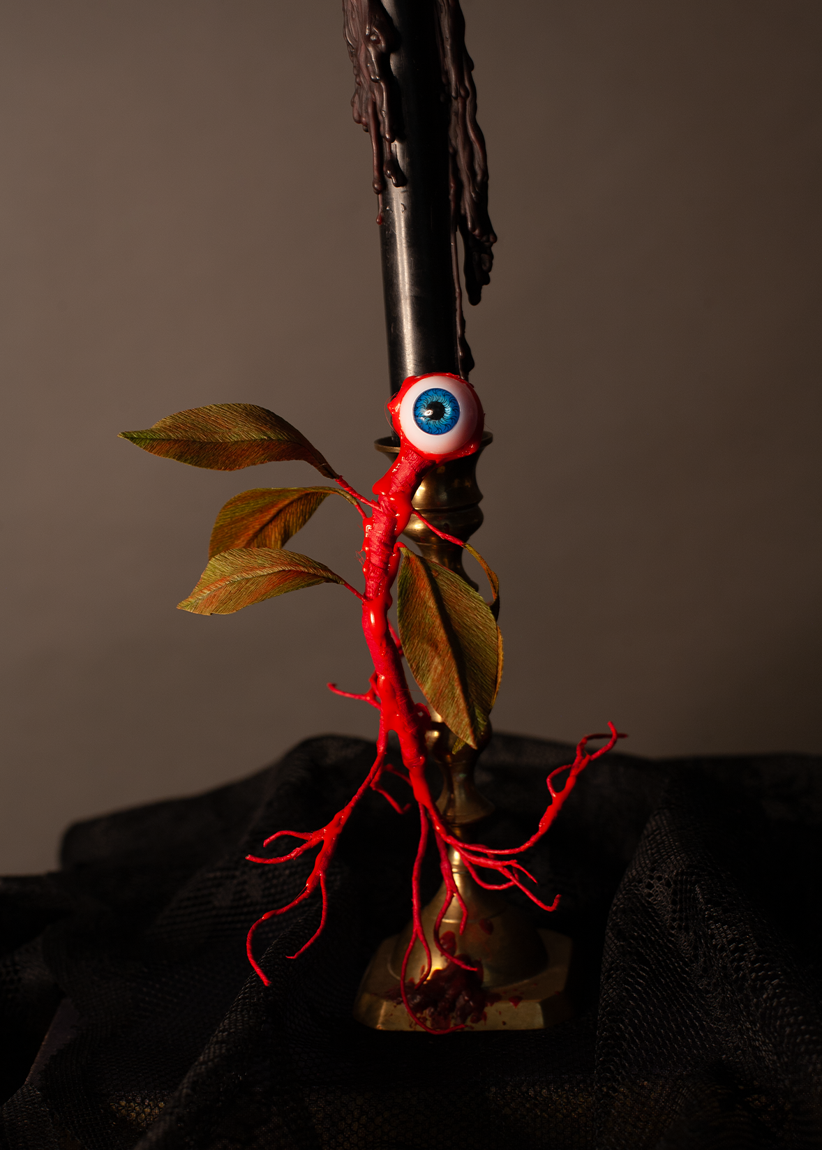 A gothic decorative object featuring a creature with a realistic red root system and a single blue-and-white eyeball, accented with brown leaves, set against a black candle and a dark draped background.