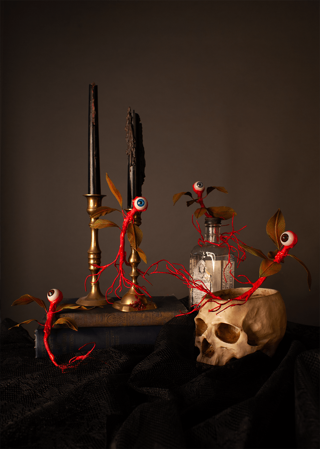 Gothic creature arrangement featuring red thread eyeball vines with green brown leaves draped around a skull, candlesticks, a glass bottle, and a dark cloth against a dark brown background.