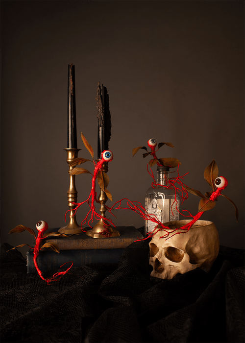Gothic creature arrangement featuring red thread eyeball vines with green brown leaves draped around a skull, candlesticks, a glass bottle, and a dark cloth against a dark brown background.