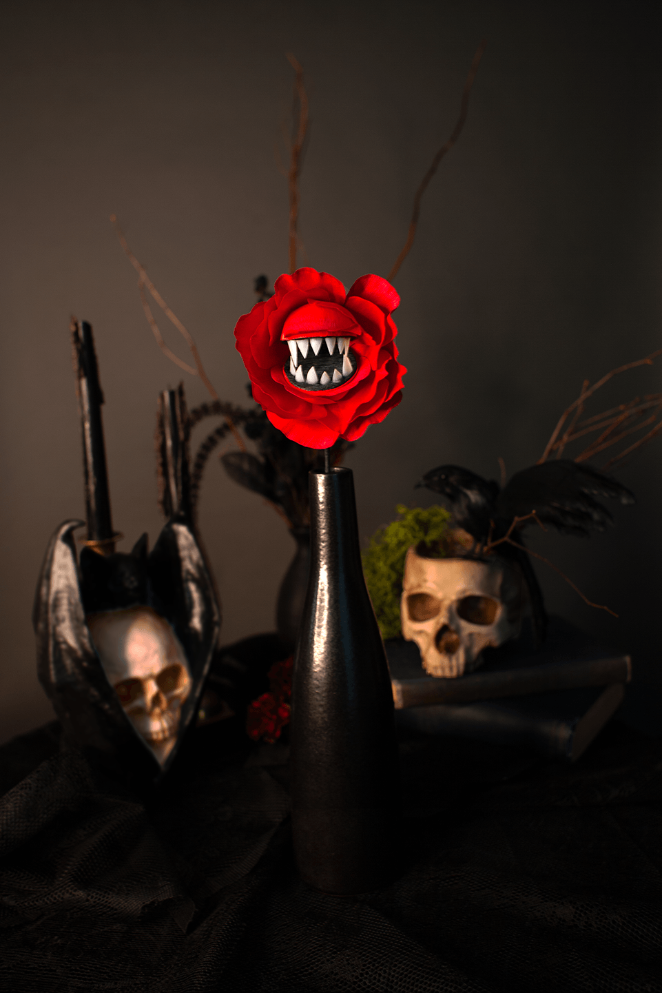 A gothic-style tabletop arrangement featuring a bright red paper rose with a fanged mouth in a black vase, flanked by skull decorations and black feathers against a dark background.