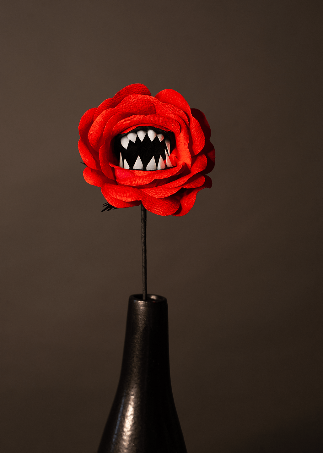 A red paper flower stem with a mouth filled with jagged white teeth in the center, displayed in a glossy black vase against a dark grey background.