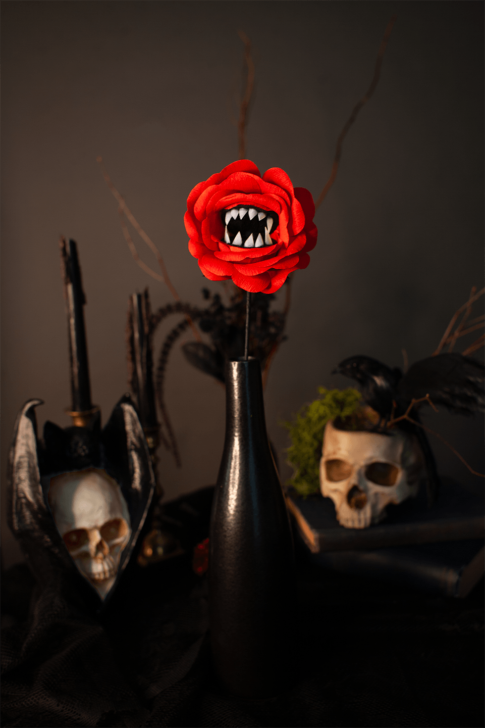 A gothic-style decorative arrangement featuring a bright red artificial paper rose with white fangs in a black vase, surrounded by skulls, black candles, and dark props against a dark background.
