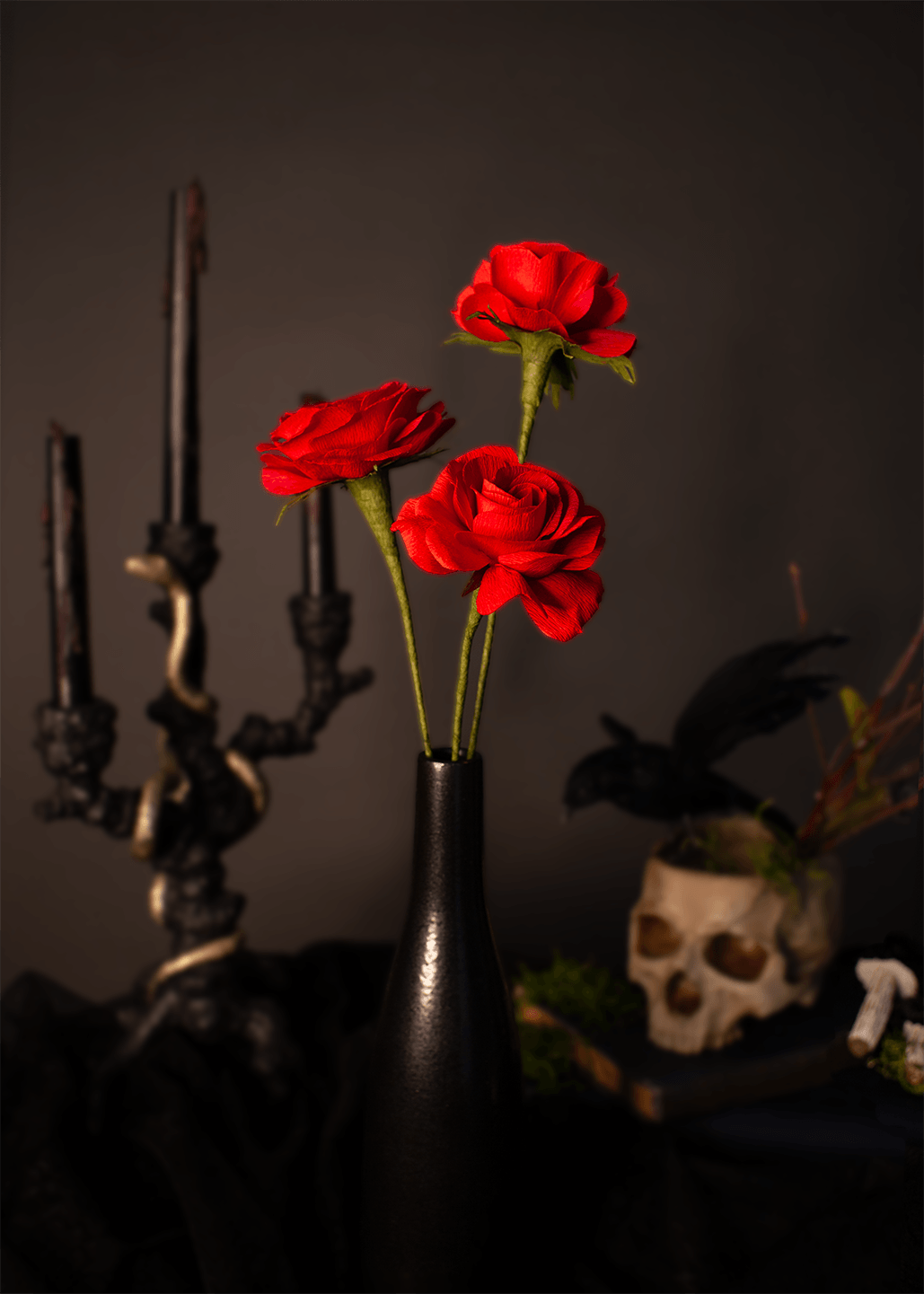 Three red paper roses in a black vase, styled with gothic props including a black candelabra and a skull against a dark background.