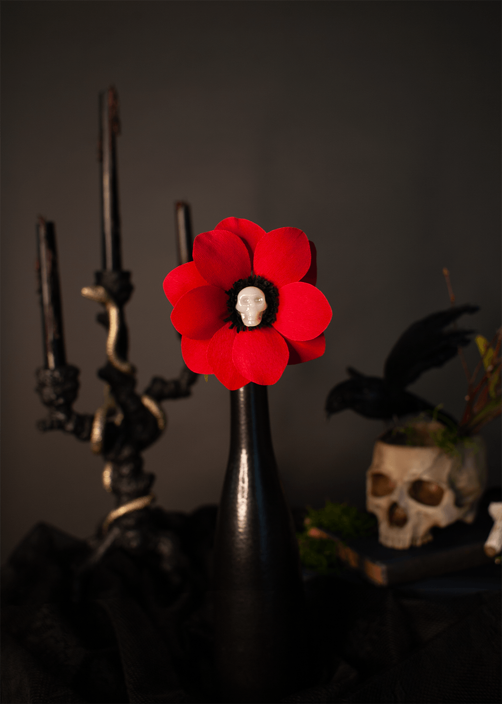 A gothic-style decorative flower arrangement featuring a vibrant red paper flower with a small white skull at its center, mounted on a glossy black vase. The vase is set against a dark, moody background with a black candelabra on the left and a skull planter on the right.