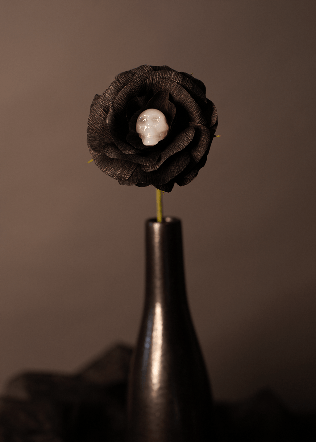 A black paper flower with a small white skull centerpiece, inserted into a dark metallic vase against a dark grey background.