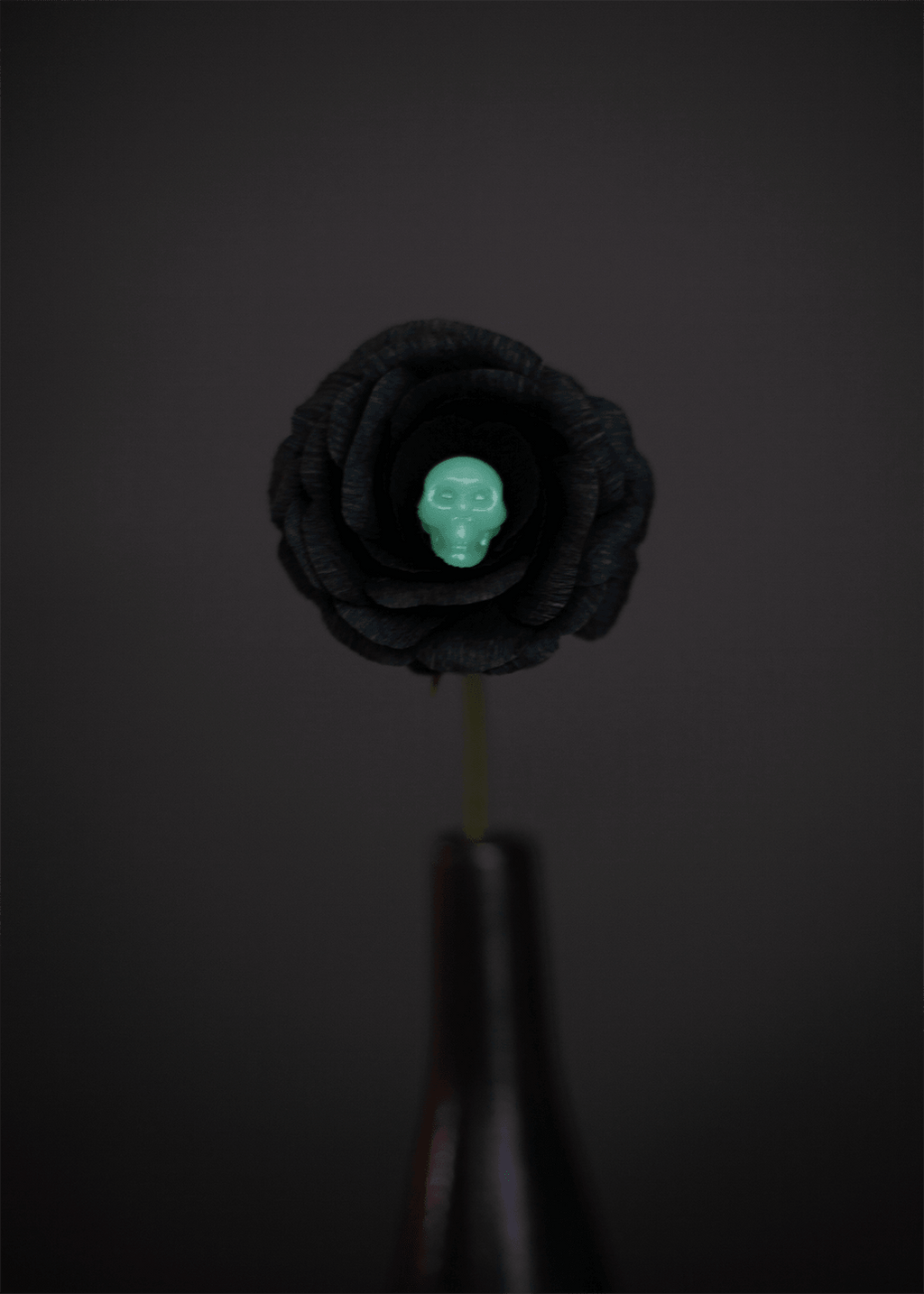 A black paper rose with a small glow-in-the-dark skull embedded in a flower at its center, mounted on a dark stem against a dark gray background.