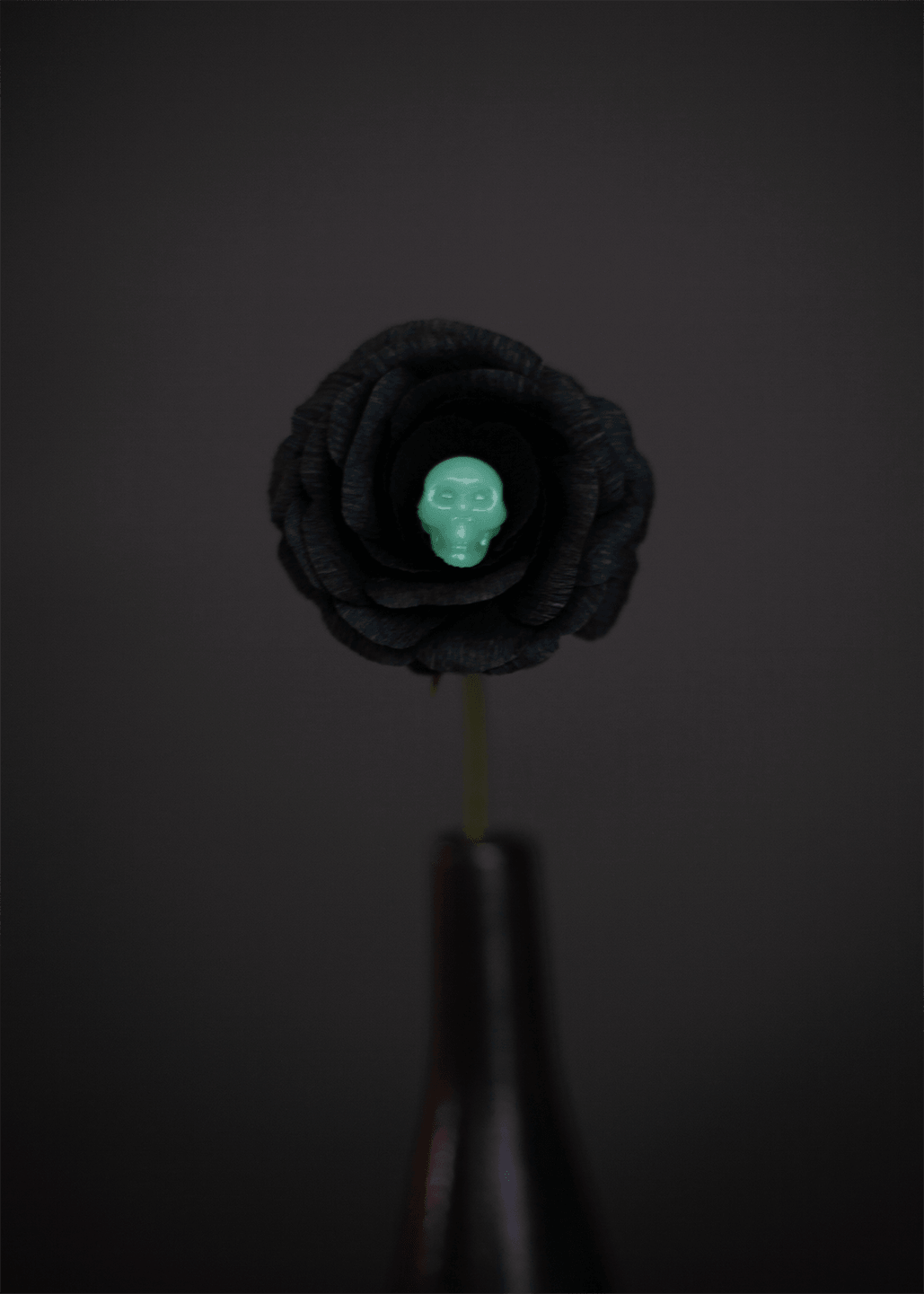 A black paper rose with a small glow-in-the-dark skull embedded in a flower at its center, mounted on a dark stem against a dark gray background.