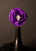 A purple paper rose with a white skull embedded in a flower at its center, mounted on a dark stem against a dark gray background.