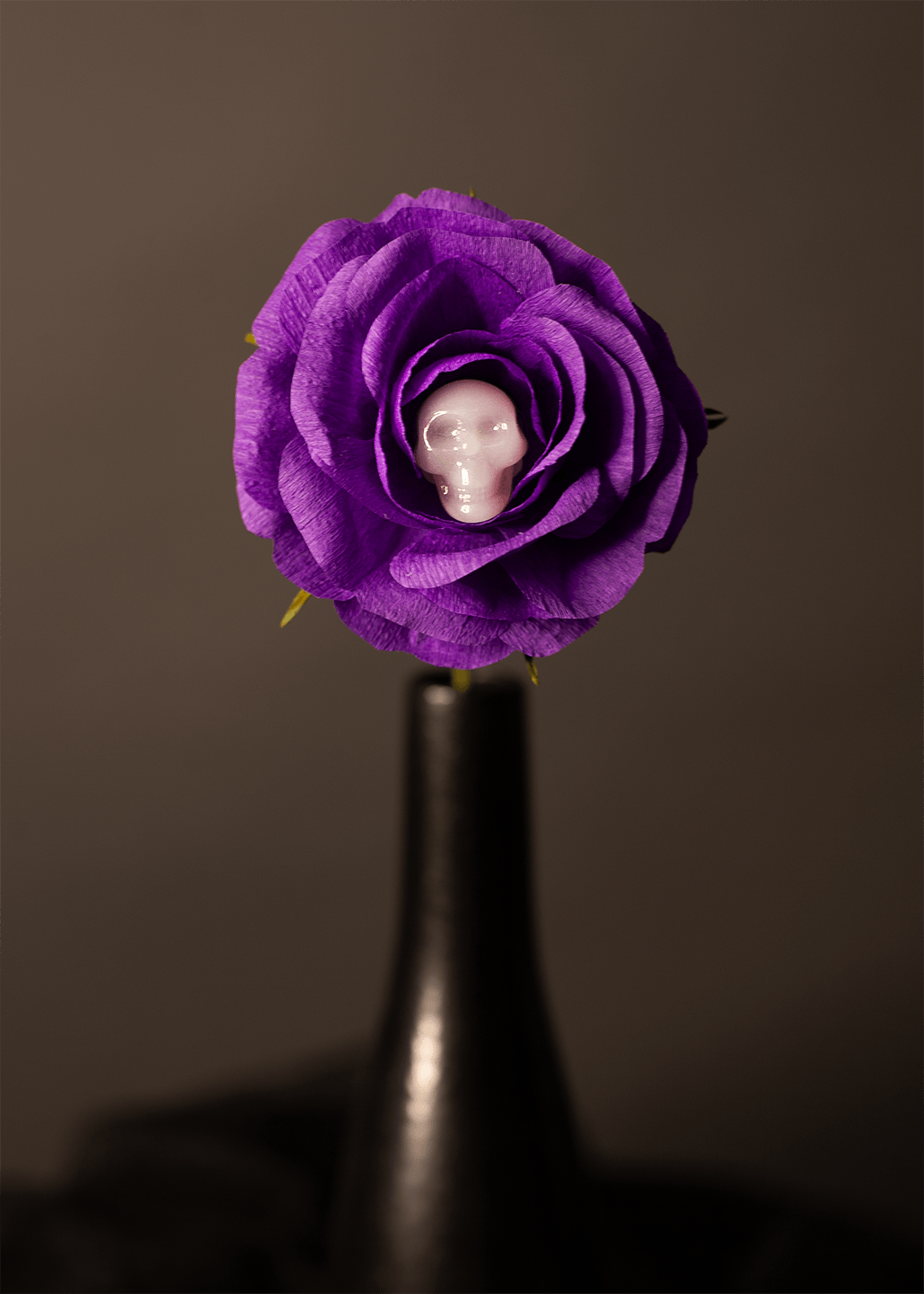 A purple paper rose with a white skull embedded in a flower at its center, mounted on a dark stem against a dark gray background.