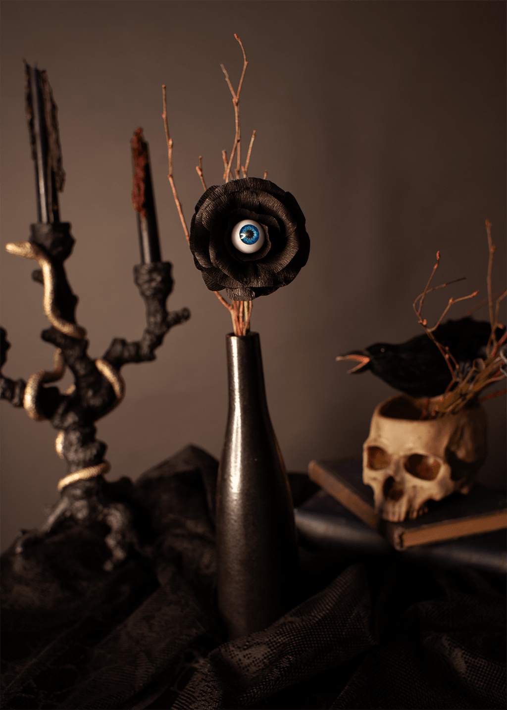A gothic-style still life featuring a black ceramic vase with a paper black rose bloom that has a realistic blue and white eye in the center, set against a dark brown background with a skull, candelabra, and crow.