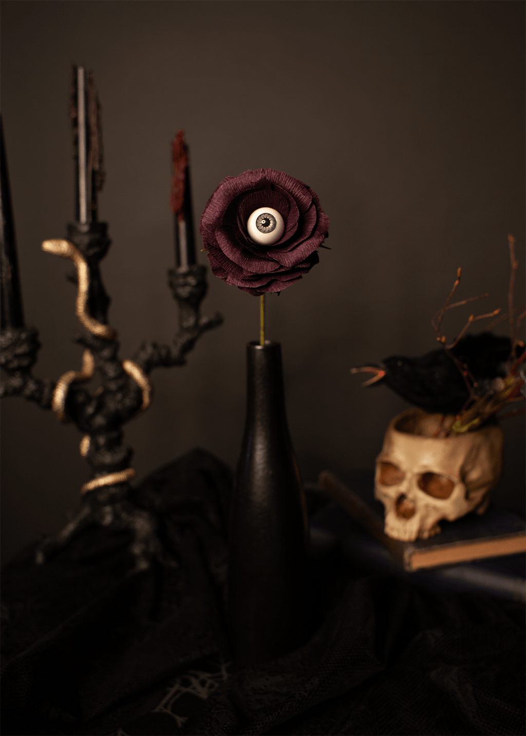 A single gothic dark purple rose with a realistic human eye at its center, inserted into a matte black vase. The vase is set on a black cloth next to a black candelabra and a skull-shaped planter.