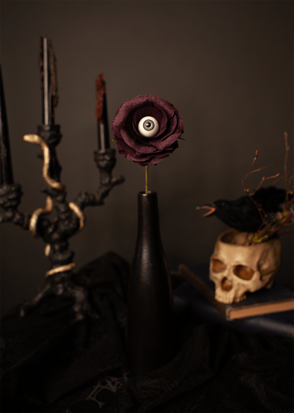 A single gothic dark purple rose with a realistic human eye at its center, inserted into a matte black vase. The vase is set on a black cloth next to a black candelabra and a skull-shaped planter.