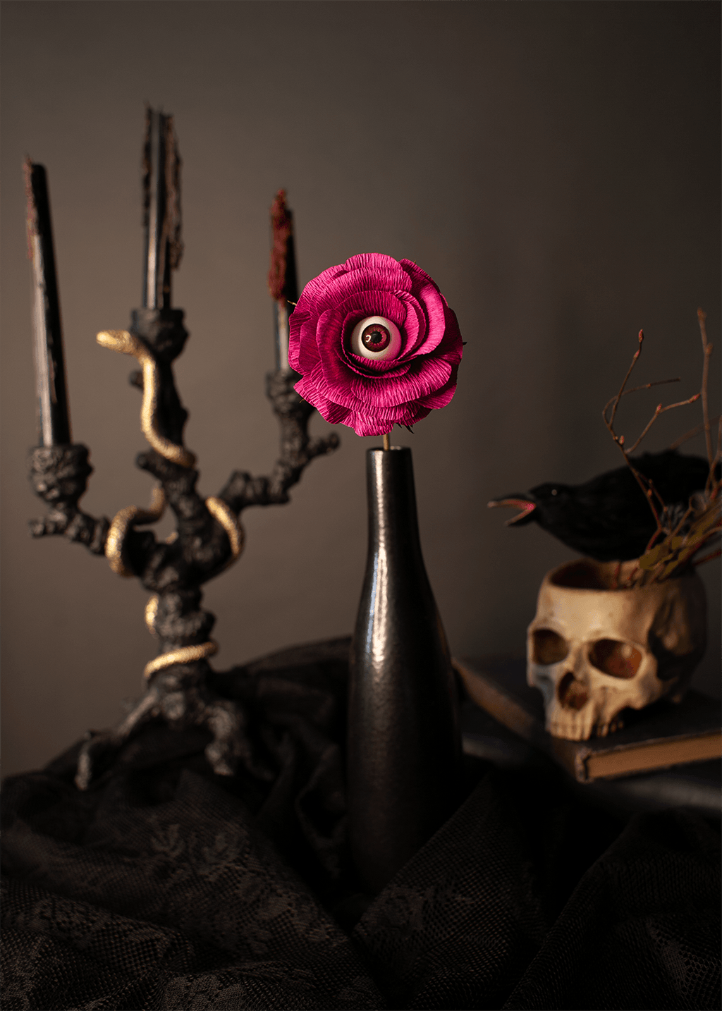 A dark, moody still life featuring a bright magenta-pink paper rose with a realistic eye in the center, held in a black bottle. The background includes a black candelabra, a skull planter with a crow, and black fabric.