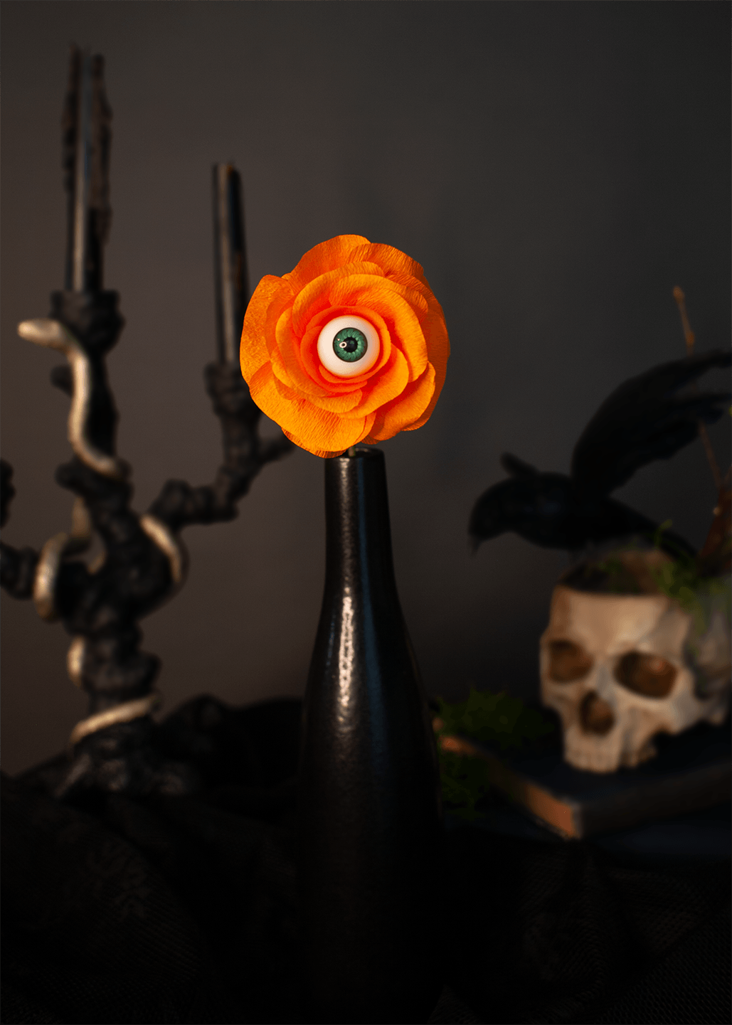 An orange paper flower with a realistic green-and-white eye in the center, displayed on a glossy black bottle against a dark gray background with gothic props including black candles, a skull, and a raven.