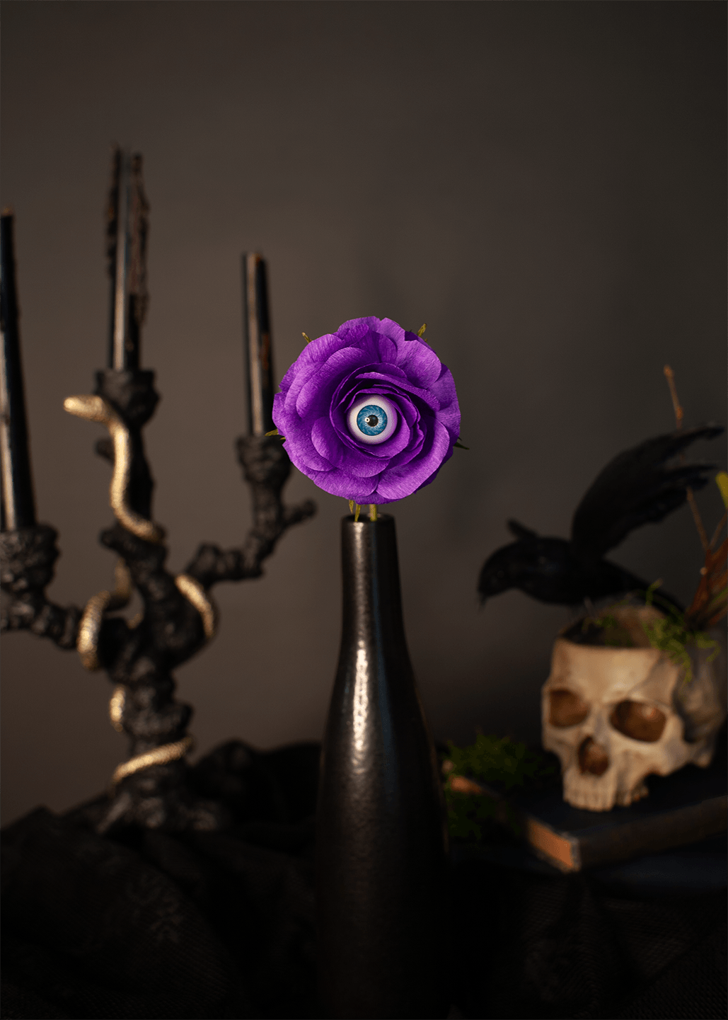 A single purple paper rose with a blue and white evil eye center, displayed in a dark glass bottle on a black cloth, styled with a black candelabra and a skull in a moody setting.