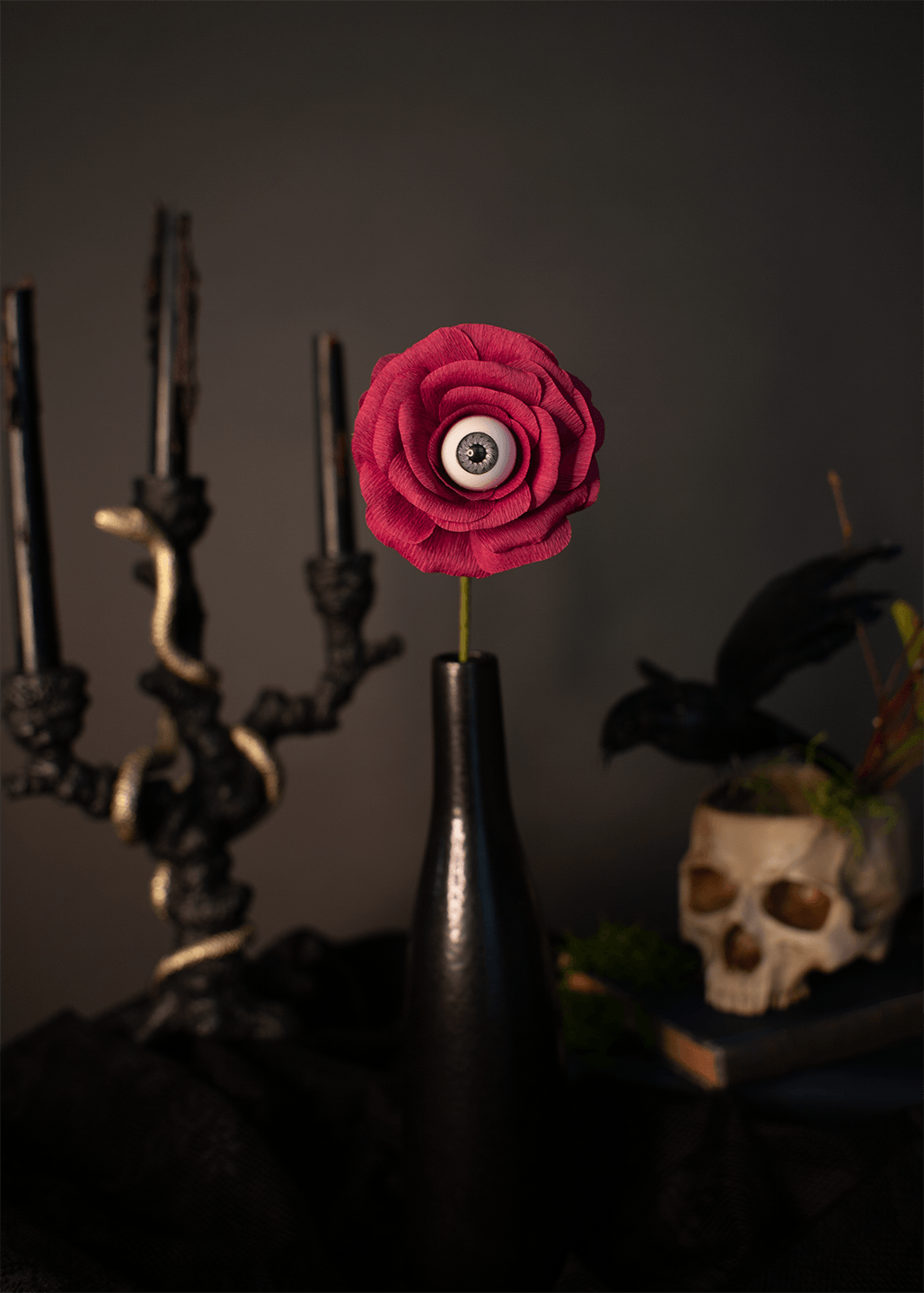 A dark, moody gothic still life featuring a single sangria-pink paper rose with a realistic human eye at its center, standing in a glossy black vase. The background is dark and atmospheric, decorated with a black candelabra, a skull, and a raven.