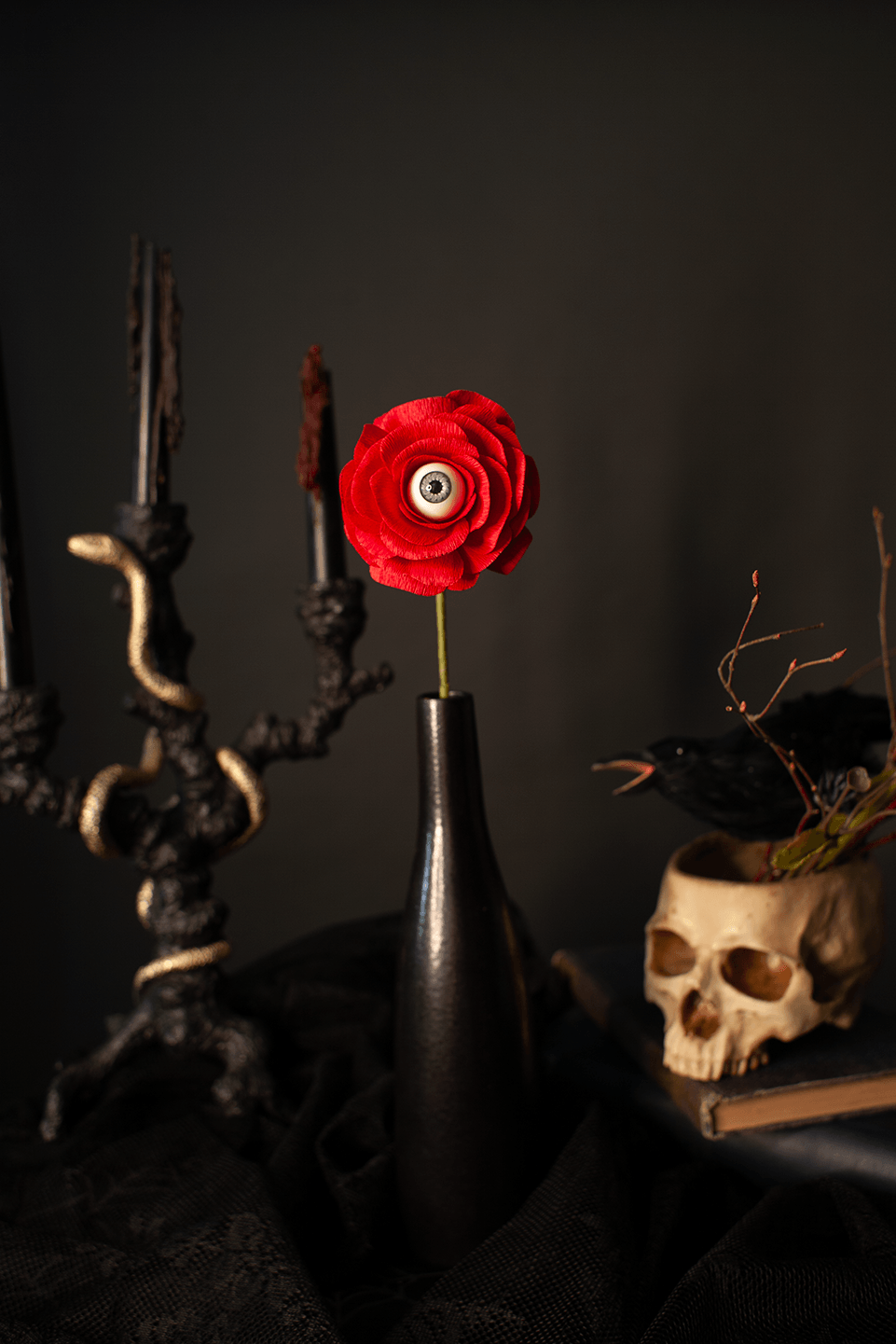 A gothic-style still life featuring a single red paper flower with a realistic eye in the center, set in a dark matte vase. The scene is styled with a black candelabra on the left and a skull-shaped planter with a black bird on the right, all against a dark gray background.