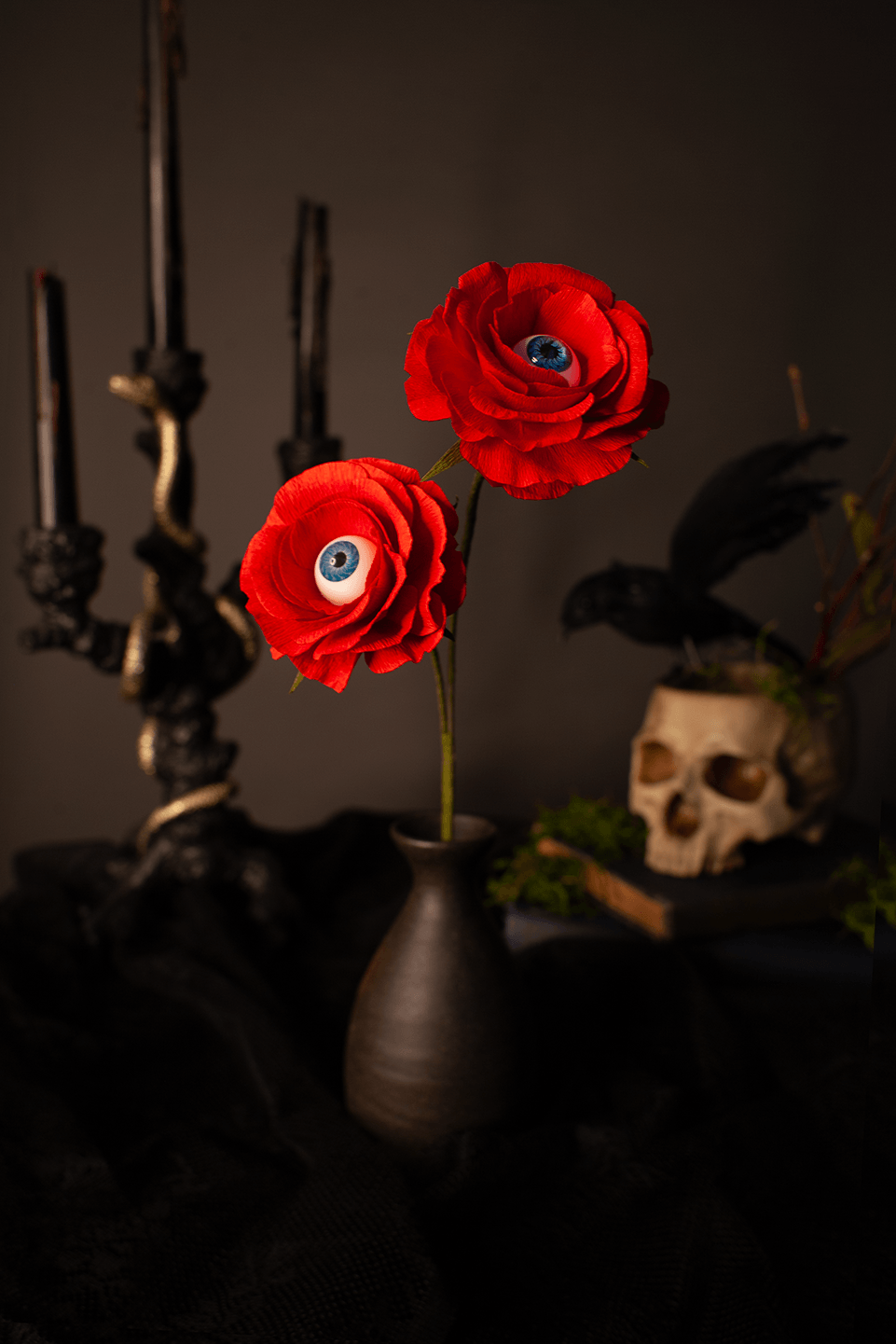 Two red artificial paper roses with realistic blue and white eyeballs in the center of the petals, arranged in a dark brown ceramic vase on a black cloth-covered surface, with gothic props including a candelabra, skull, and crow in the background.